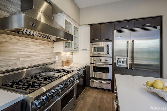 kitchen with a warming drawer, tasteful backsplash, glass insert cabinets, built in appliances, and wall chimney exhaust hood