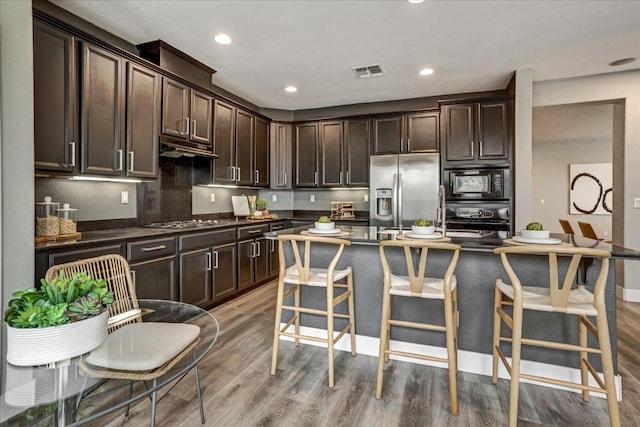 kitchen featuring appliances with stainless steel finishes, hardwood / wood-style floors, a kitchen bar, dark brown cabinetry, and a center island with sink