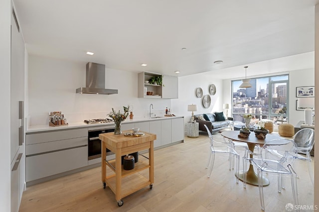 kitchen with wall oven, light wood-style floors, stainless steel gas stovetop, wall chimney exhaust hood, and a sink