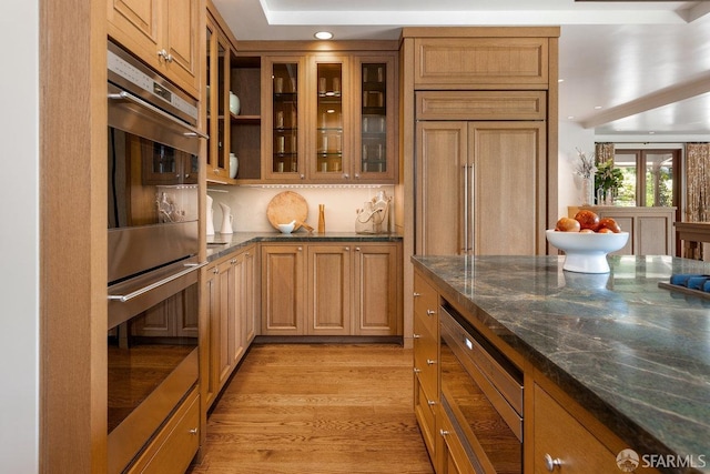 kitchen with glass insert cabinets, recessed lighting, light wood finished floors, dark stone counters, and stainless steel double oven