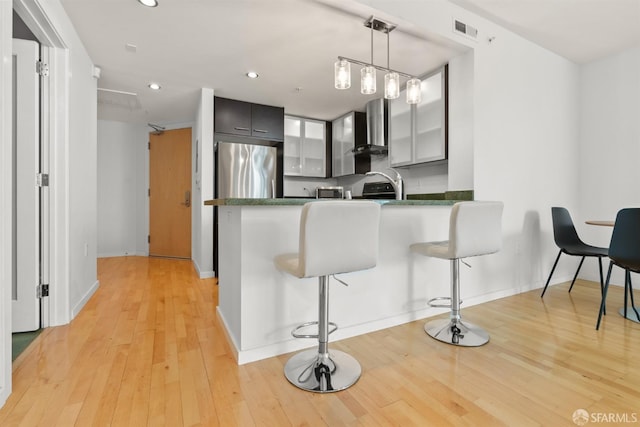 kitchen with light wood-type flooring, kitchen peninsula, stainless steel fridge, wall chimney exhaust hood, and pendant lighting