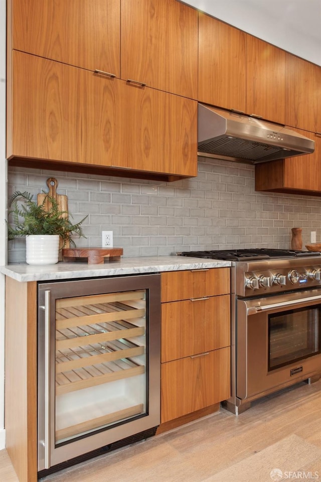 kitchen featuring beverage cooler, high end range, decorative backsplash, light stone countertops, and light wood-type flooring