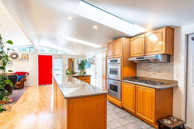 kitchen featuring lofted ceiling with skylight, under cabinet range hood, tasteful backsplash, a kitchen island, and appliances with stainless steel finishes