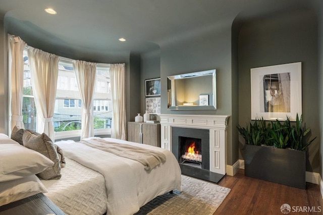 bedroom with wood finished floors, baseboards, a warm lit fireplace, and recessed lighting