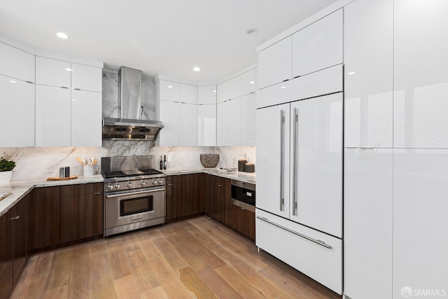 kitchen with white cabinetry, light wood-type flooring, high quality appliances, dark brown cabinetry, and wall chimney range hood