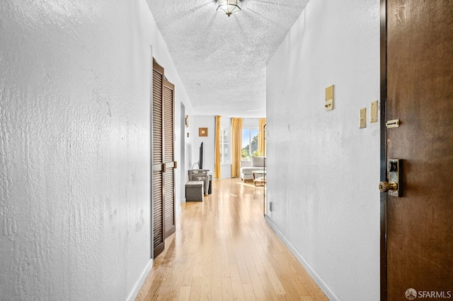 hall with baseboards, light wood-type flooring, a textured wall, and a textured ceiling