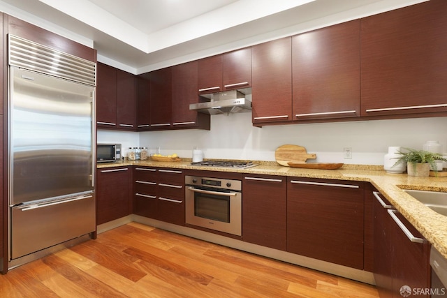 kitchen with modern cabinets, light wood-style flooring, under cabinet range hood, stainless steel appliances, and light stone countertops