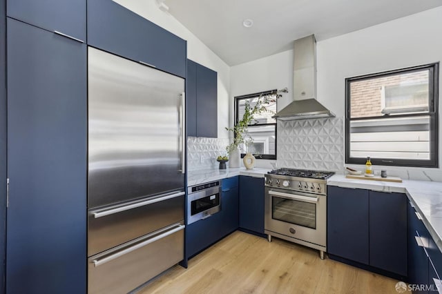 kitchen with wall chimney range hood, blue cabinetry, backsplash, light hardwood / wood-style flooring, and high quality appliances