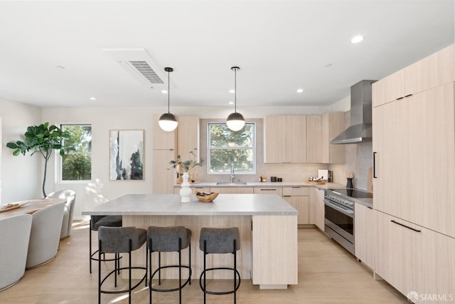 kitchen featuring light brown cabinetry, a sink, wall chimney range hood, modern cabinets, and a kitchen bar