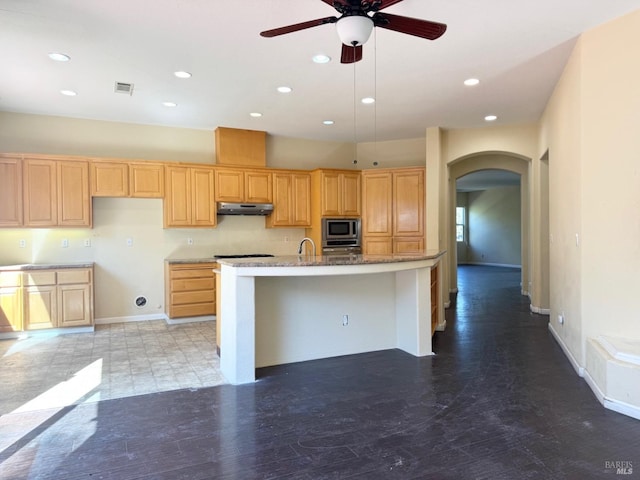 kitchen with arched walkways, stainless steel microwave, light brown cabinets, a kitchen island with sink, and under cabinet range hood