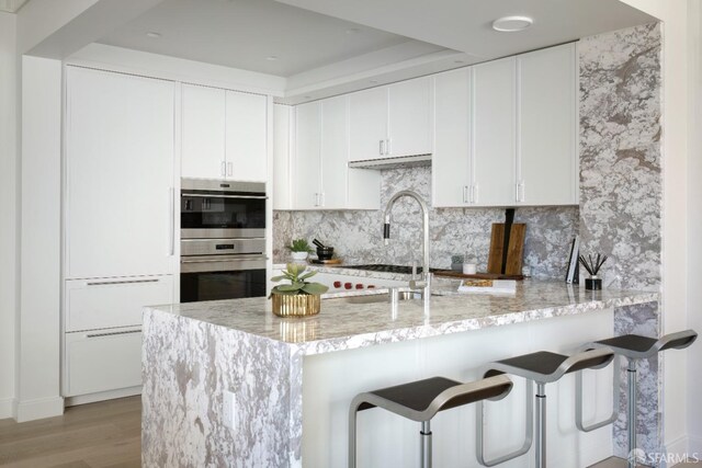 kitchen featuring stainless steel double oven, kitchen peninsula, white cabinetry, and light stone counters