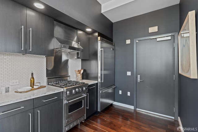 kitchen with wall chimney range hood, high end appliances, light stone counters, tasteful backsplash, and dark hardwood / wood-style flooring