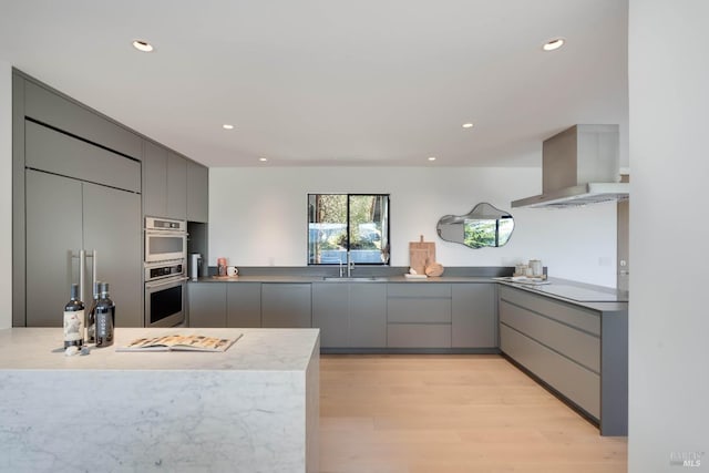 kitchen with light wood-type flooring, wall chimney range hood, gray cabinetry, stainless steel double oven, and modern cabinets