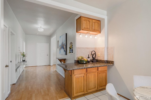 kitchen with brown cabinets, decorative backsplash, dark stone countertops, light wood-type flooring, and a peninsula