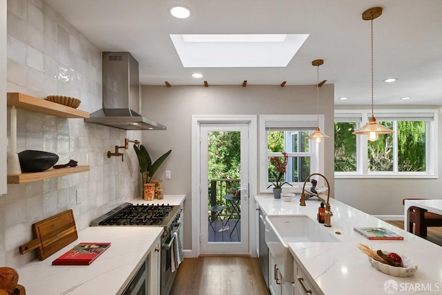 kitchen featuring a sink, appliances with stainless steel finishes, a skylight, wall chimney range hood, and light stone countertops