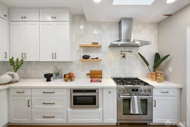 kitchen featuring light stone countertops, appliances with stainless steel finishes, white cabinetry, wall chimney exhaust hood, and tasteful backsplash