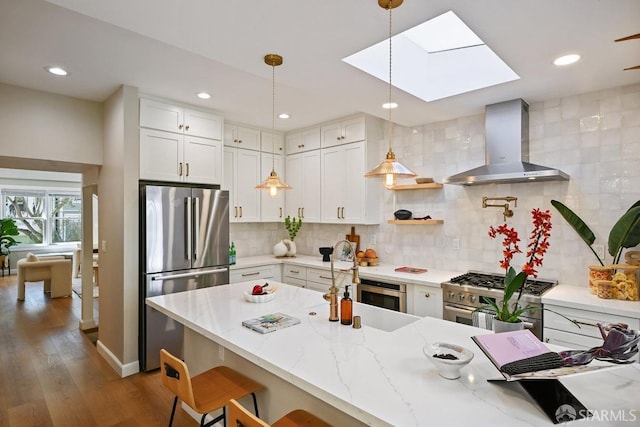 kitchen with a breakfast bar area, a skylight, appliances with stainless steel finishes, pendant lighting, and wall chimney range hood