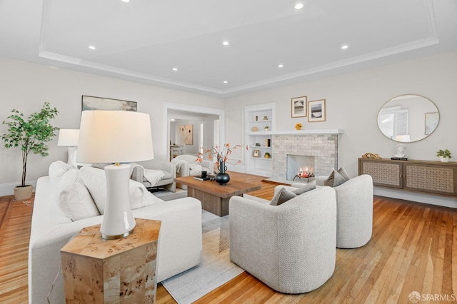 living room with a brick fireplace, ornamental molding, a tray ceiling, built in features, and light hardwood / wood-style floors