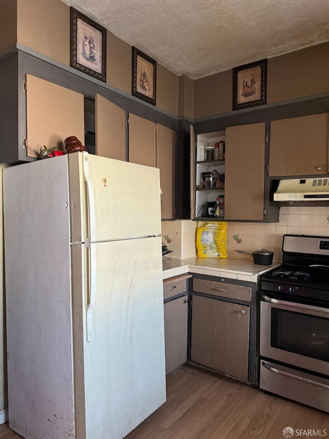 kitchen with white refrigerator, stainless steel range with gas cooktop, tile countertops, range hood, and light hardwood / wood-style floors