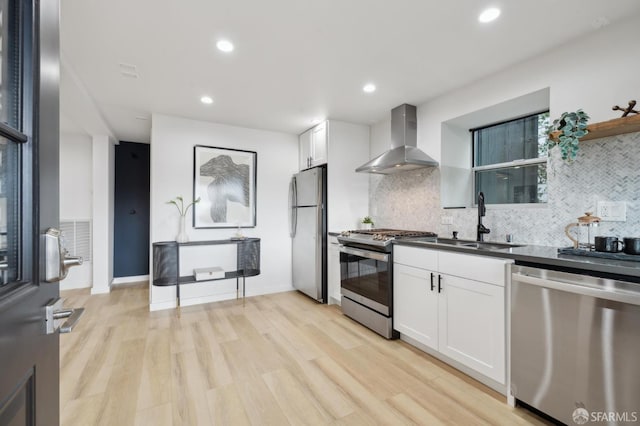 kitchen featuring light wood-type flooring, a sink, appliances with stainless steel finishes, white cabinets, and wall chimney range hood