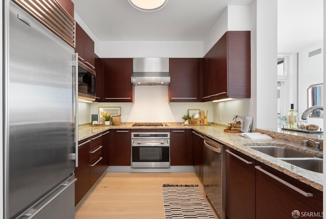 kitchen featuring wall chimney range hood, light stone counters, a sink, light wood-style floors, and built in appliances
