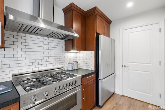 kitchen with appliances with stainless steel finishes, backsplash, wall chimney exhaust hood, brown cabinetry, and dark countertops