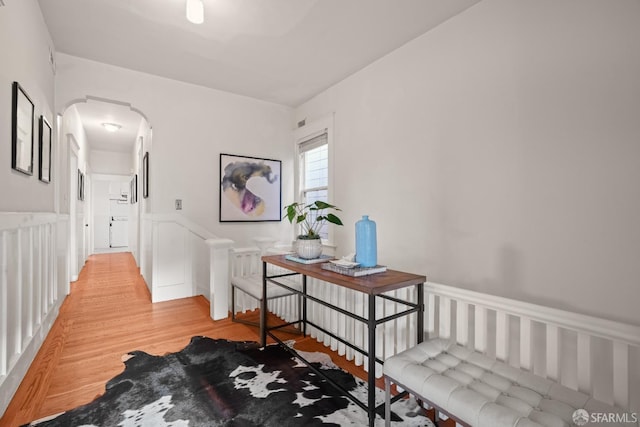 hall featuring arched walkways, light wood finished floors, a wainscoted wall, and a decorative wall