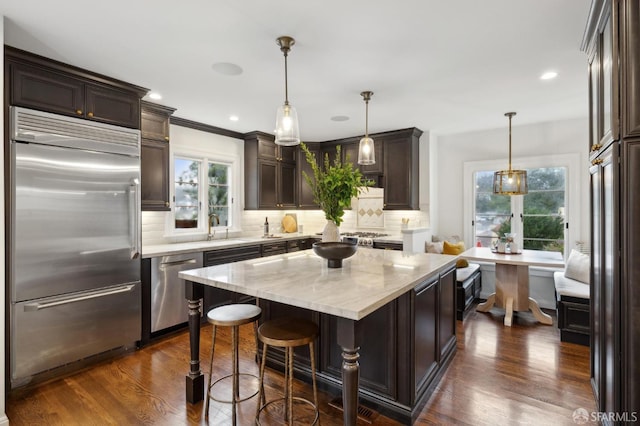 kitchen featuring appliances with stainless steel finishes, hanging light fixtures, a center island, dark brown cabinetry, and light stone countertops