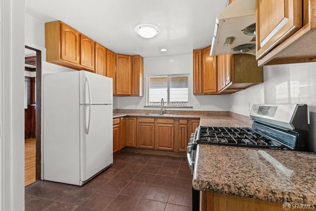 kitchen featuring stainless steel gas stove, brown cabinetry, freestanding refrigerator, range hood, and a sink