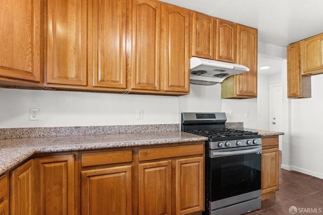 kitchen with baseboards, brown cabinets, under cabinet range hood, dark tile patterned floors, and stainless steel range with gas stovetop