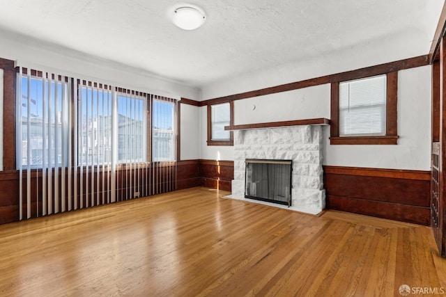 unfurnished living room featuring a textured ceiling, a stone fireplace, a wainscoted wall, and wood finished floors