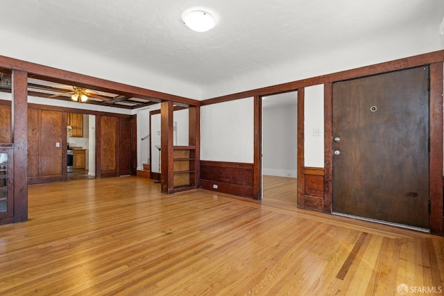 unfurnished living room with light wood-style floors, a ceiling fan, a wainscoted wall, and beamed ceiling