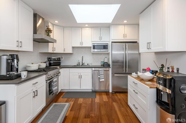 kitchen with a skylight, a sink, stainless steel appliances, dark countertops, and wall chimney exhaust hood