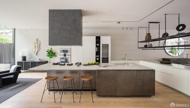 kitchen with white cabinets, sink, an island with sink, wall oven, and light hardwood / wood-style floors