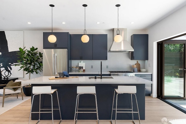 kitchen with an island with sink, sink, decorative light fixtures, high end fridge, and wall chimney range hood