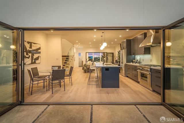 kitchen featuring hanging light fixtures, a center island with sink, wall chimney exhaust hood, appliances with stainless steel finishes, and a breakfast bar area