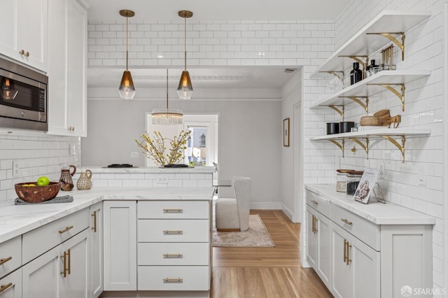 kitchen with stainless steel microwave, decorative light fixtures, white cabinetry, light stone countertops, and light wood-type flooring