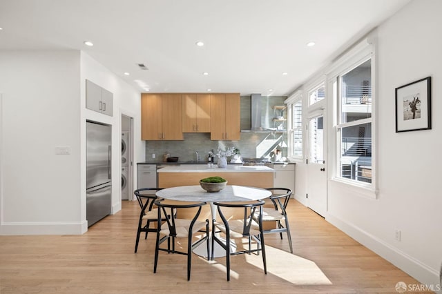 kitchen with wall chimney exhaust hood, stacked washer / drying machine, tasteful backsplash, light wood-type flooring, and stainless steel built in fridge