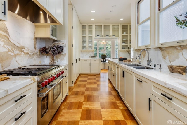 kitchen with light stone counters, a sink, range hood, range with two ovens, and glass insert cabinets