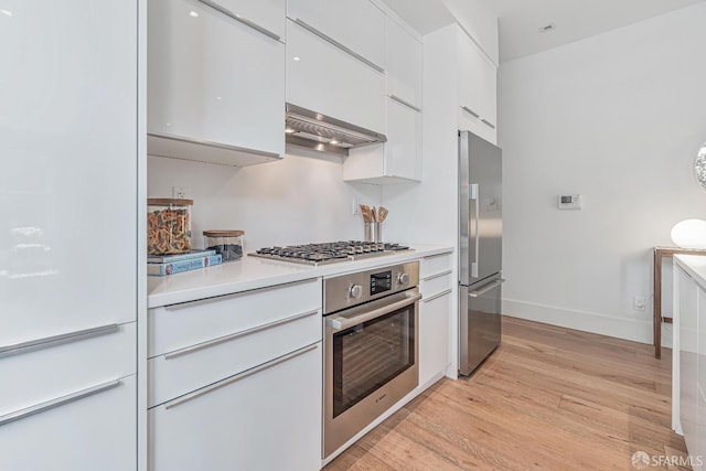 kitchen with light countertops, appliances with stainless steel finishes, light wood-style floors, white cabinets, and wall chimney exhaust hood