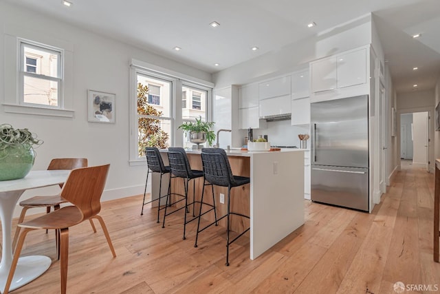 kitchen featuring modern cabinets, a center island with sink, stainless steel built in refrigerator, white cabinetry, and light wood-style floors