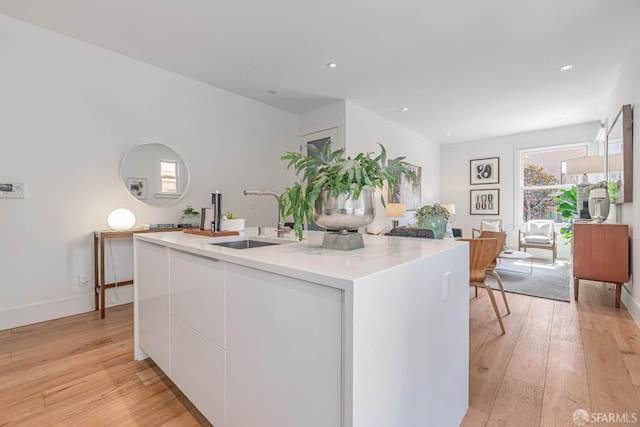 kitchen with light wood-style flooring, a center island with sink, a sink, white cabinetry, and light countertops