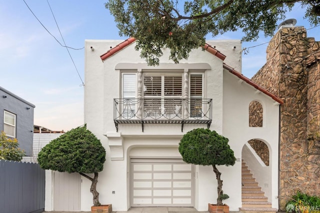 mediterranean / spanish-style house with a balcony, a garage, a tile roof, fence, and stucco siding