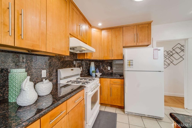 kitchen with light tile patterned floors, backsplash, dark stone counters, white appliances, and under cabinet range hood