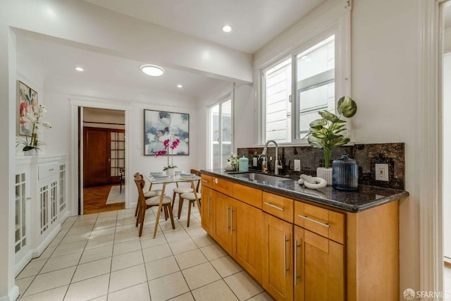 kitchen featuring light tile patterned floors, a sink, backsplash, dark stone counters, and brown cabinetry