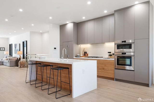 kitchen with a breakfast bar area, gray cabinetry, a kitchen island with sink, light hardwood / wood-style floors, and stainless steel double oven