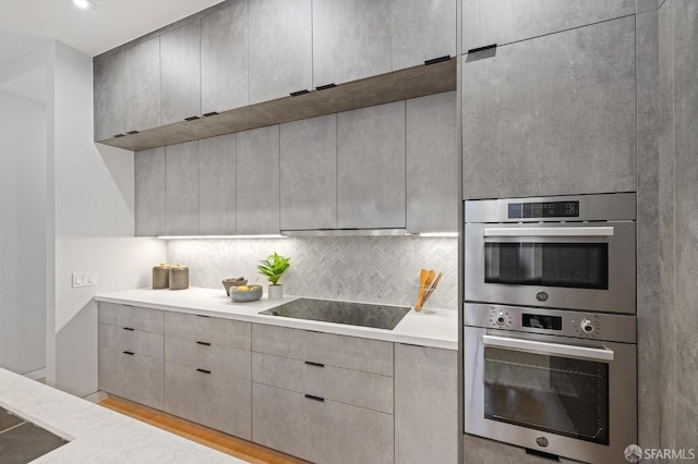 kitchen with gray cabinetry, tasteful backsplash, light wood-type flooring, stainless steel double oven, and black electric stovetop