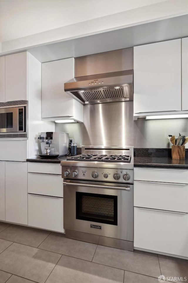 kitchen featuring wall chimney exhaust hood, dark stone countertops, appliances with stainless steel finishes, light tile patterned flooring, and white cabinetry