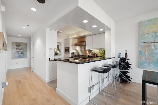 kitchen featuring kitchen peninsula, light hardwood / wood-style flooring, wall chimney exhaust hood, a kitchen bar, and white cabinetry