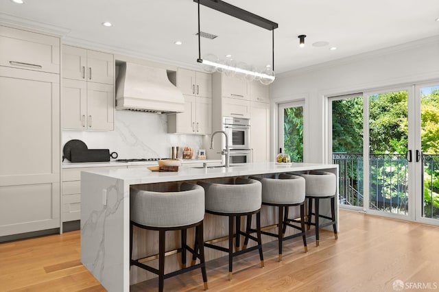 kitchen with light wood-type flooring, premium range hood, white cabinetry, and an island with sink